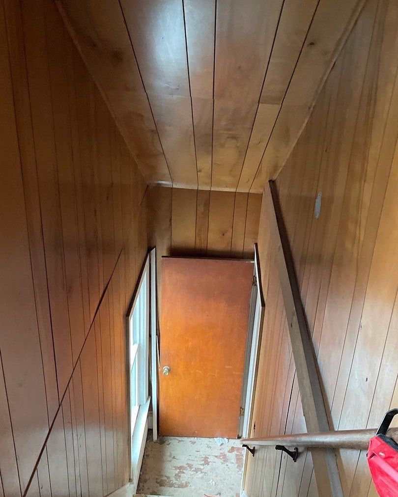Narrow hallway with wood paneling on walls and ceiling, leading to a closed brown door. Stairs and handrail on the right.