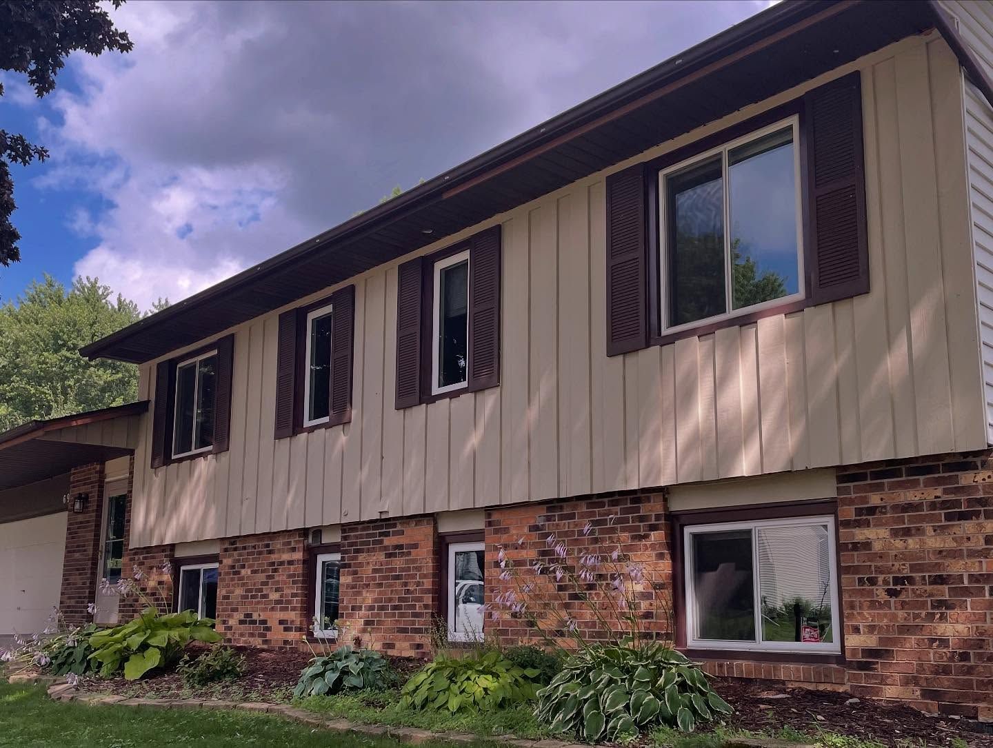 Two-story house with tan siding, brown shutters, and a brick base, under a cloudy sky.