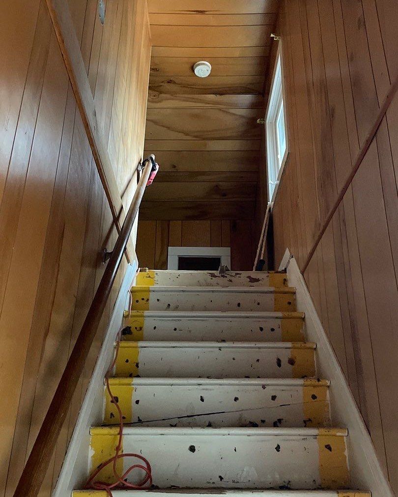 Wooden staircase with worn yellow and white steps, leading upwards. Brown paneling lines the walls, with a small window at the top.