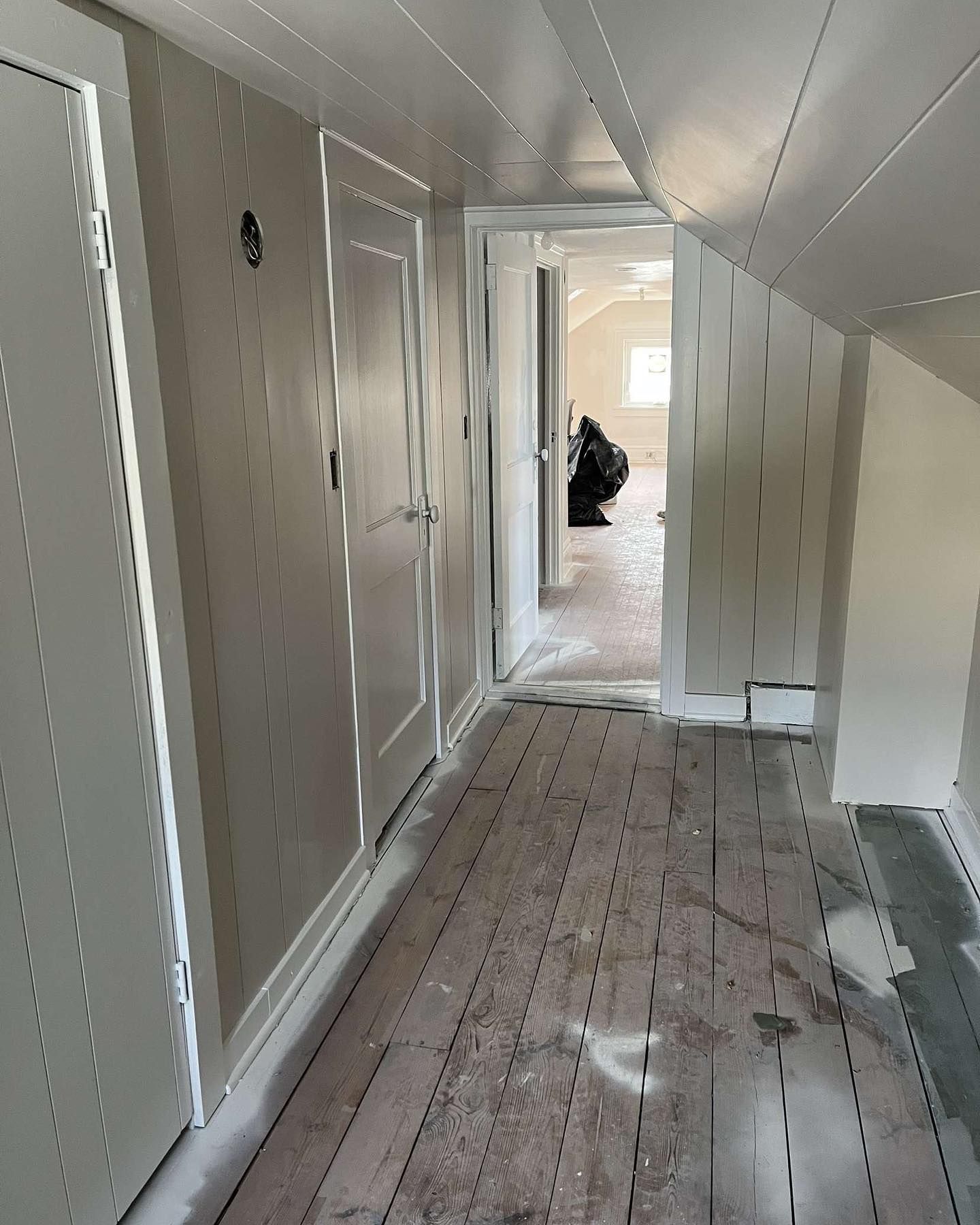 Narrow hallway with white-painted walls, doors, and ceiling, leading to a room.  Bare wooden floor with visible markings, sunlight at the end.