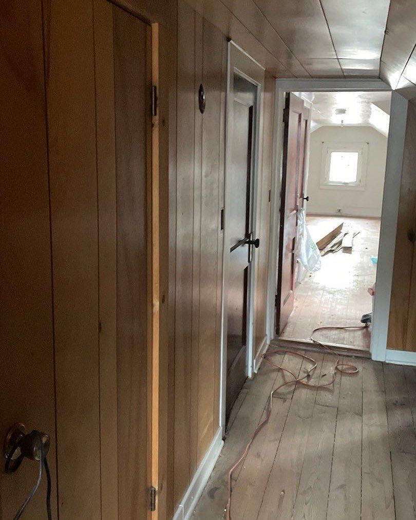 Narrow hallway with wooden walls and doors, leading to a room with a window and construction materials.
