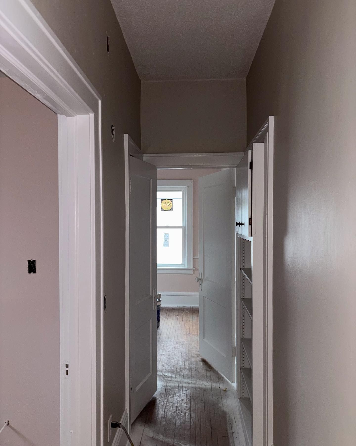 A hallway with white trim and doors, beige walls, and wooden floors leading to a window at the end.