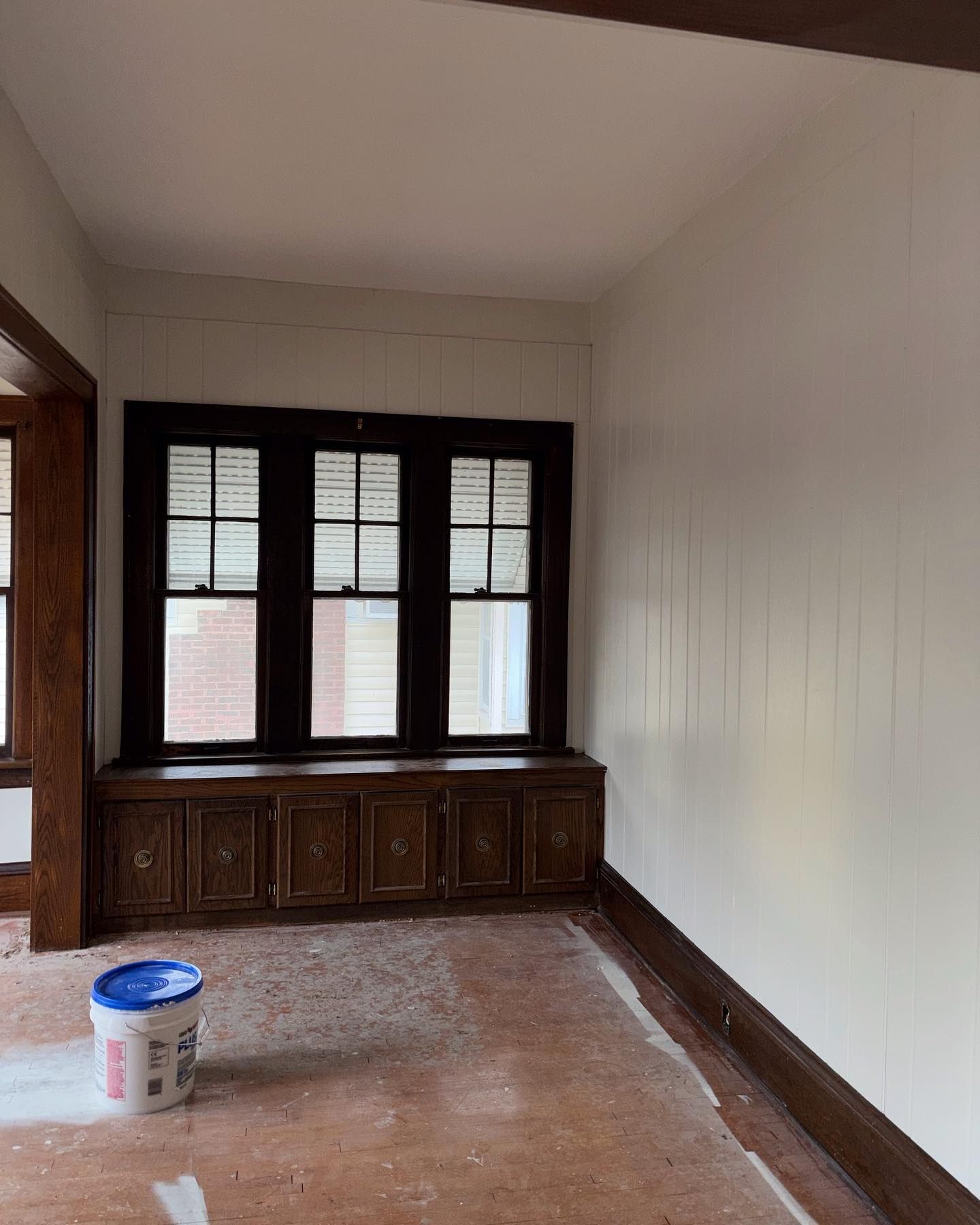 Interior of a room with dark-stained trim, white walls, and a window with a built-in cabinet. A paint bucket sits on the floor.