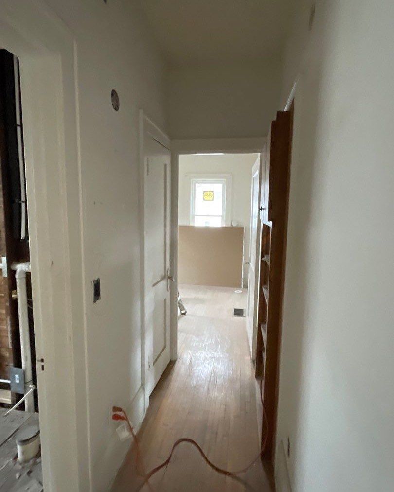 Narrow hallway with white walls and wood flooring. A door is on the left and a doorway leads to another room, suggesting renovation.