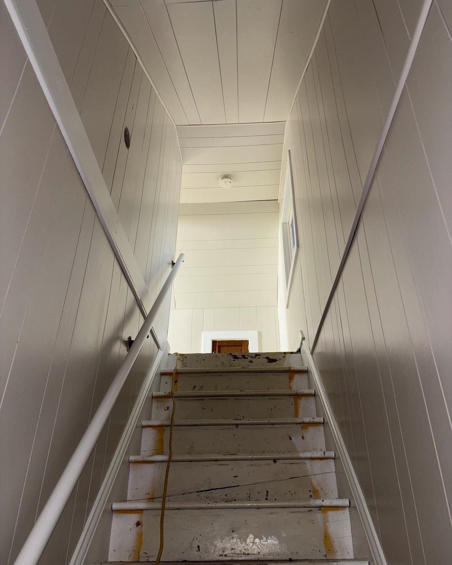 Staircase leading upwards with white walls and handrails. Worn wooden steps with a view of a doorway at the top.