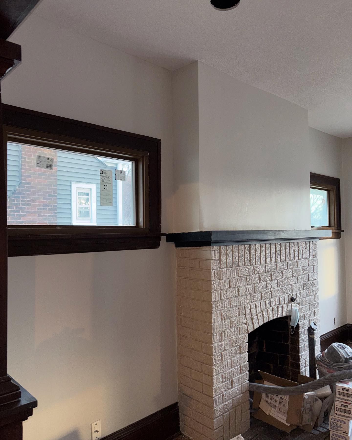 Interior with a brick fireplace, two windows with dark brown trim, and white walls. The windows look out onto a neighboring house.