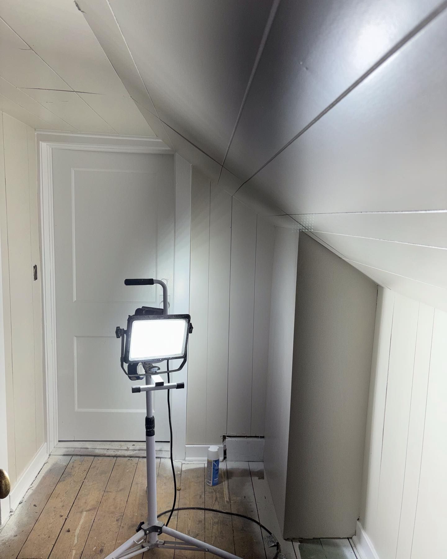 Brightly lit, freshly painted attic room with a closed white door, angled walls, and a work light on a tripod.