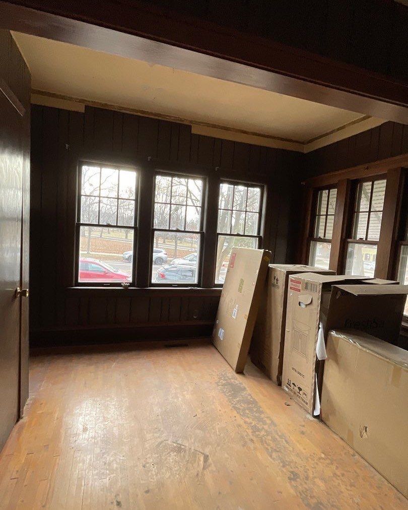 Room interior with wooden paneling, windows, and cardboard boxes. A door is on the left, and sunlight shines through the windows.