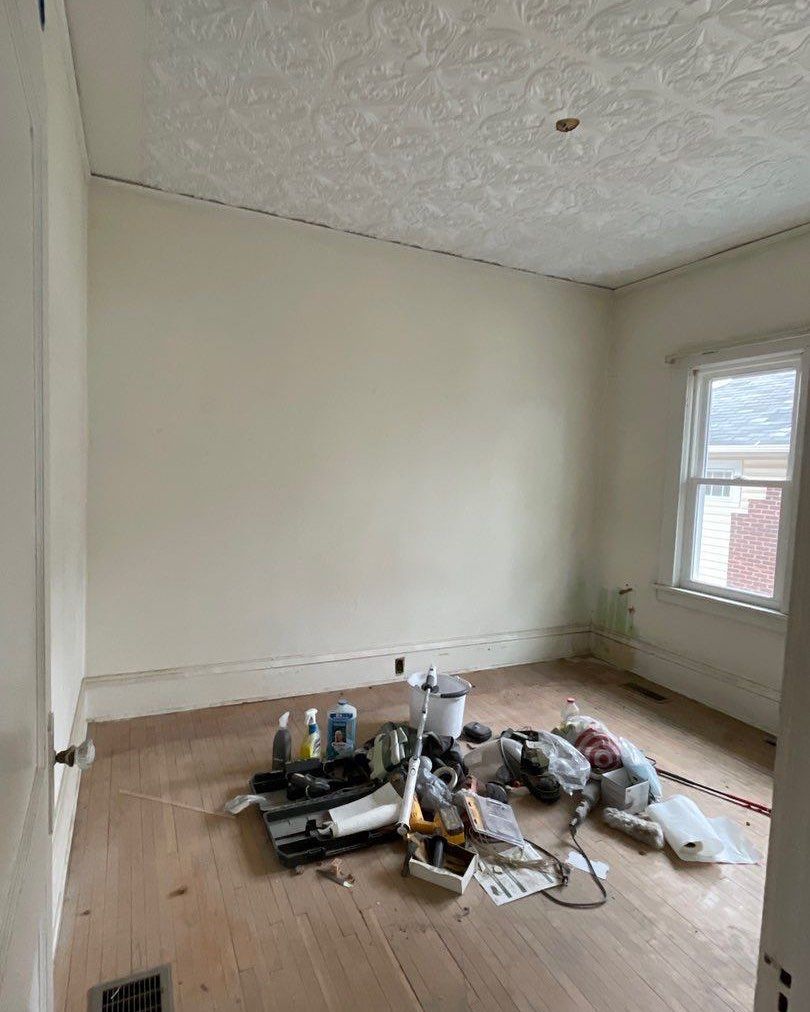 Empty room with bare wood floors, freshly painted cream walls, and a window. Construction supplies are piled on the floor.