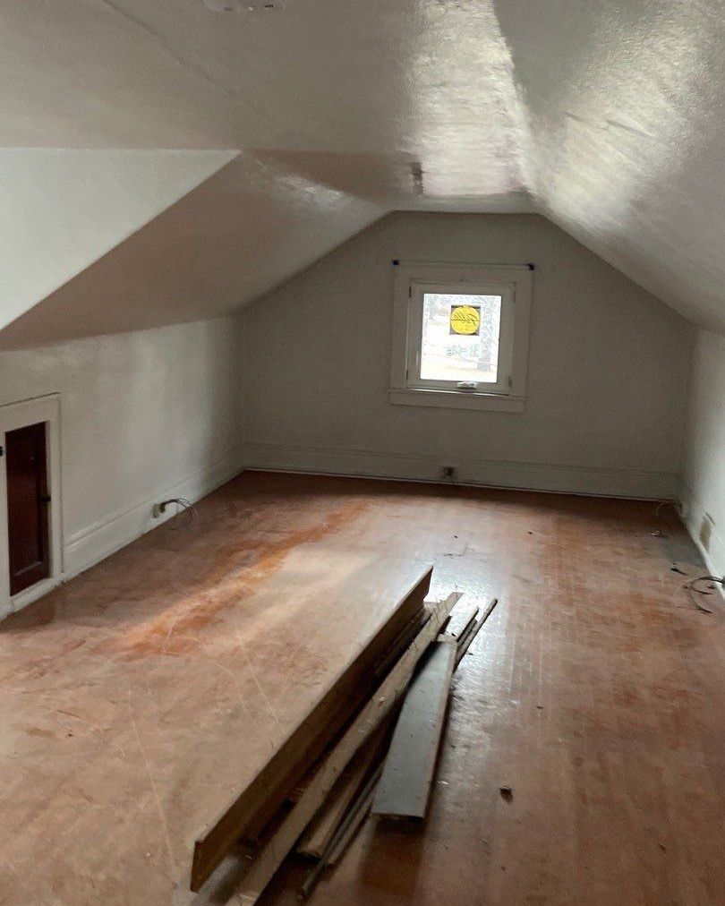 Attic room with angled ceilings, a small window, and weathered wooden floor. Boards lie on the floor.