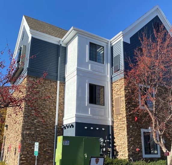 Exterior view of a three-story building with blue siding, stone accents, and a white center section. Red and orange fall foliage is in the foreground.