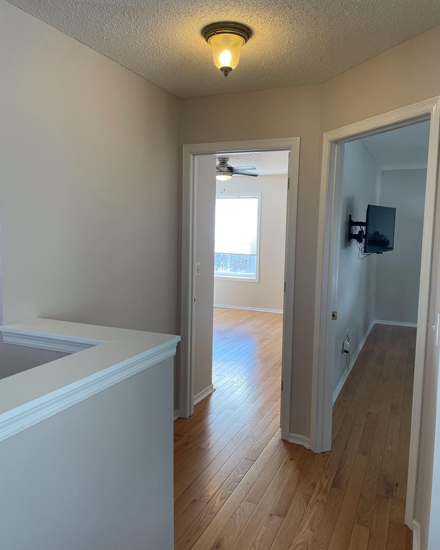 Hallway with hardwood floors and three doorways, one leading to a room with a window, another to a room with a mounted TV.