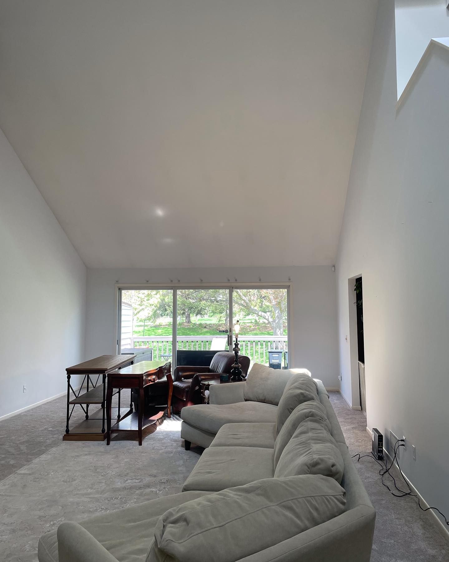 Spacious living room with a high, angled ceiling, light-colored sofa, desk, and sliding glass doors overlooking greenery.