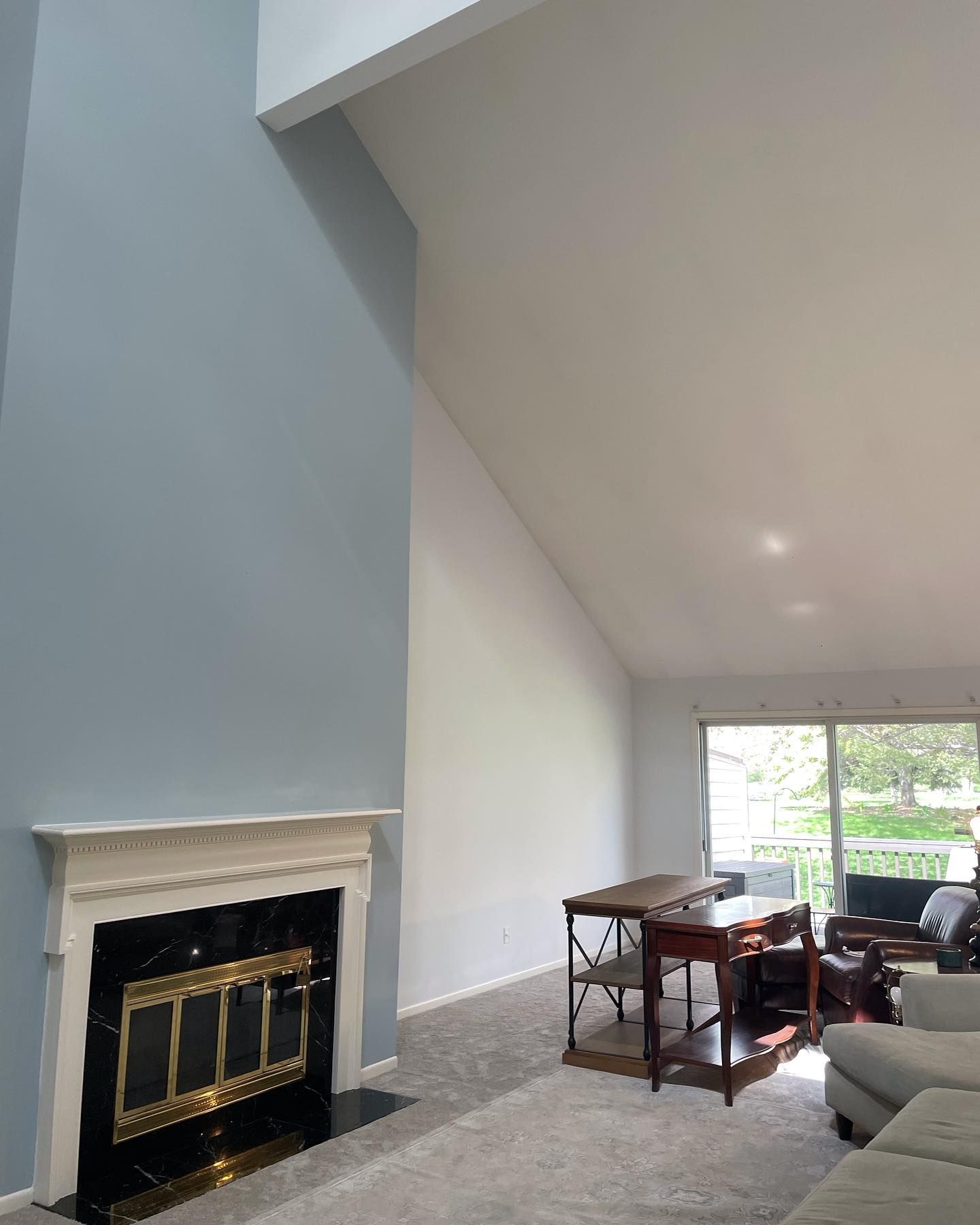 A living room with a fireplace, high ceilings, and a light blue wall. There's a table, couch, and sliding glass door to an outside view.