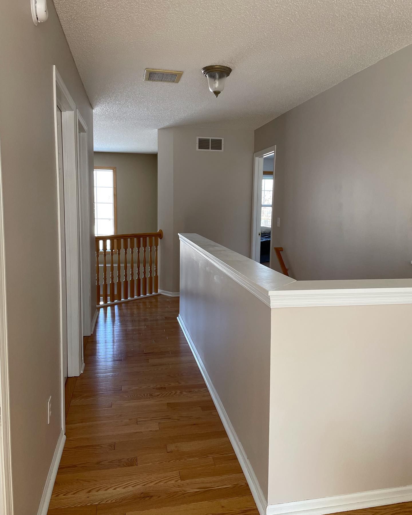 Hallway with hardwood floors, beige walls, and a white-trimmed balcony railing. Light enters from a window at the end.