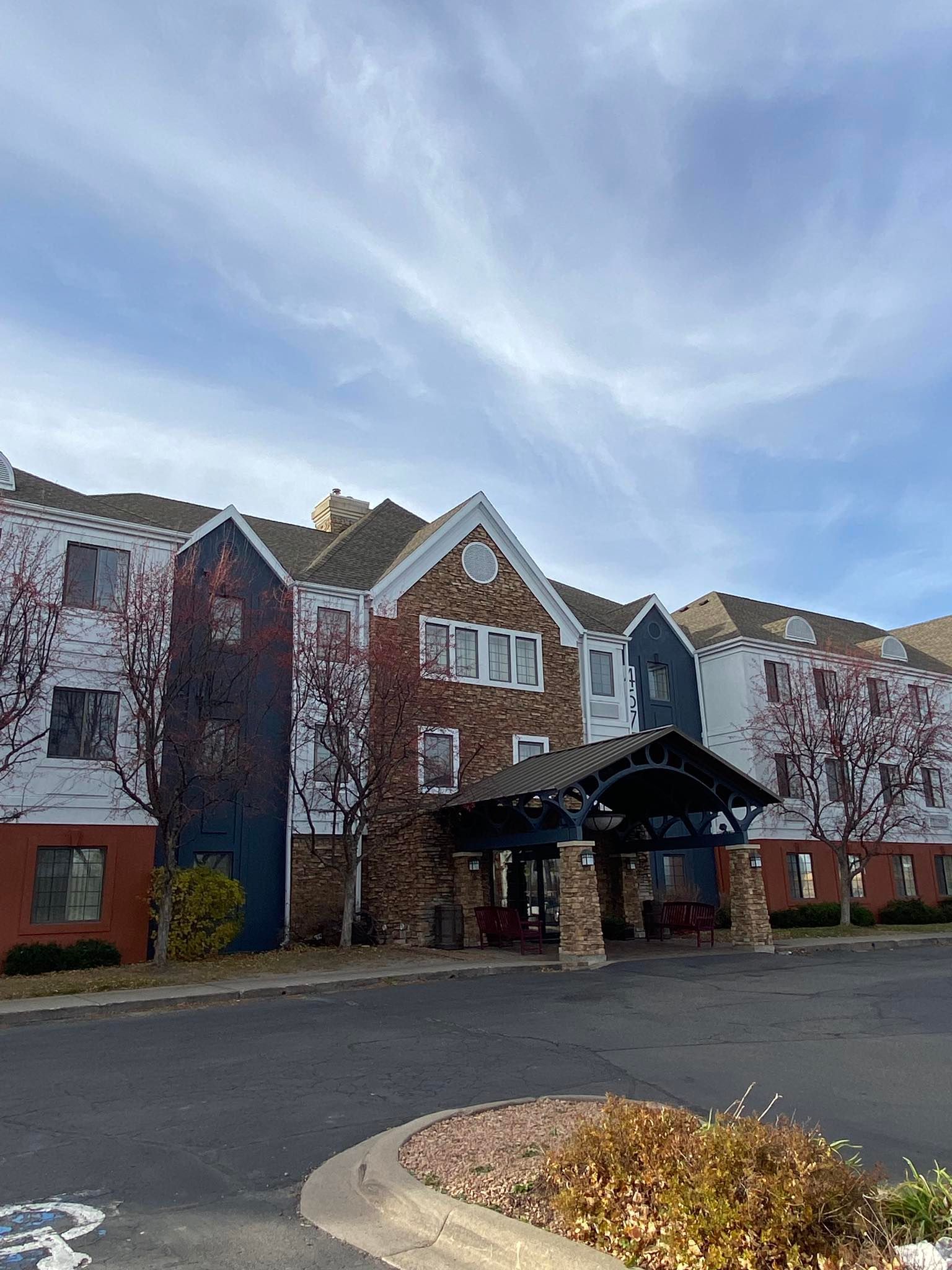Hotel exterior with stone and white siding under a cloudy sky. Entrance has a covered canopy. Dry vegetation is visible.