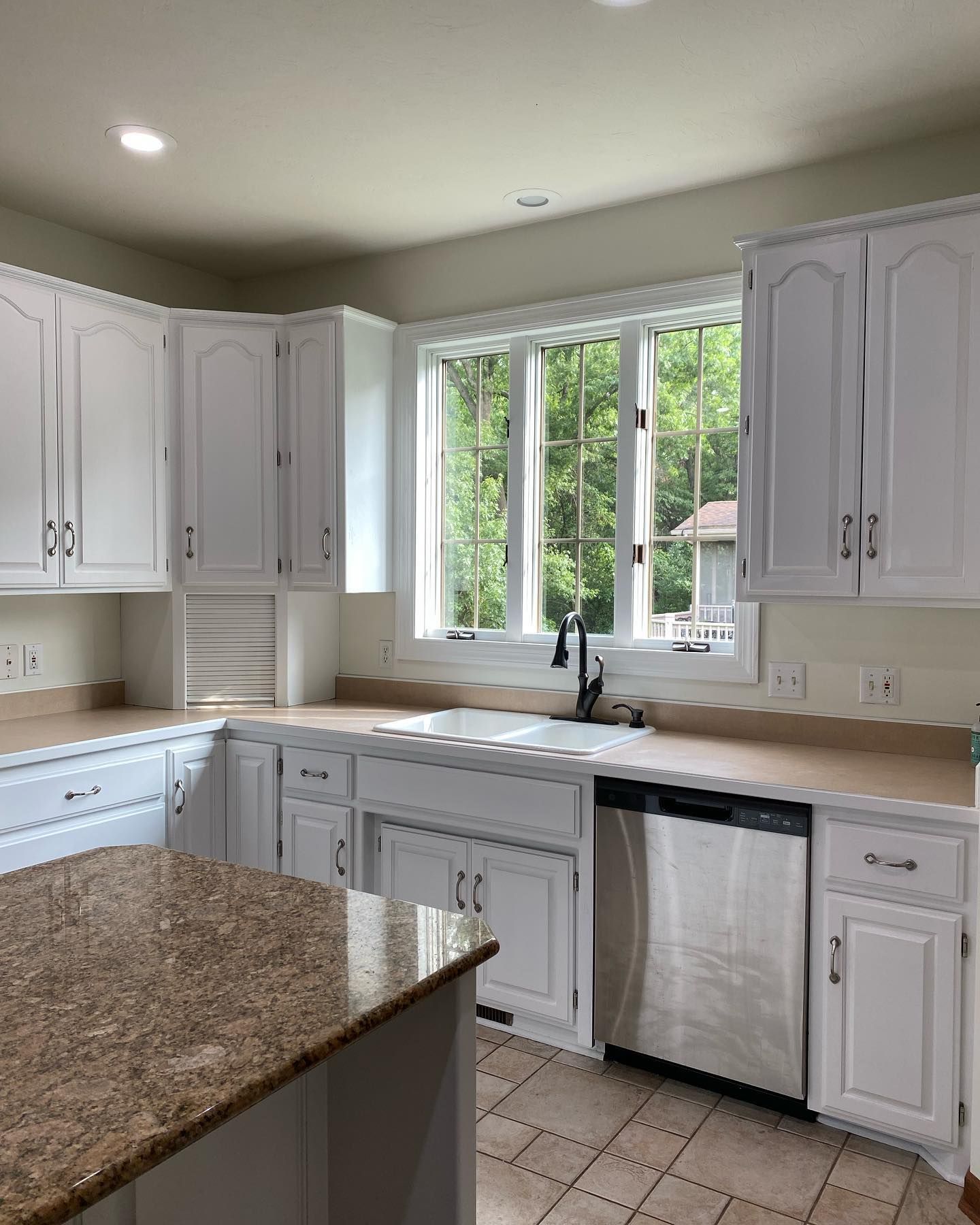 White kitchen with granite countertops and stainless steel appliances. A window overlooks a lush green outdoor setting.