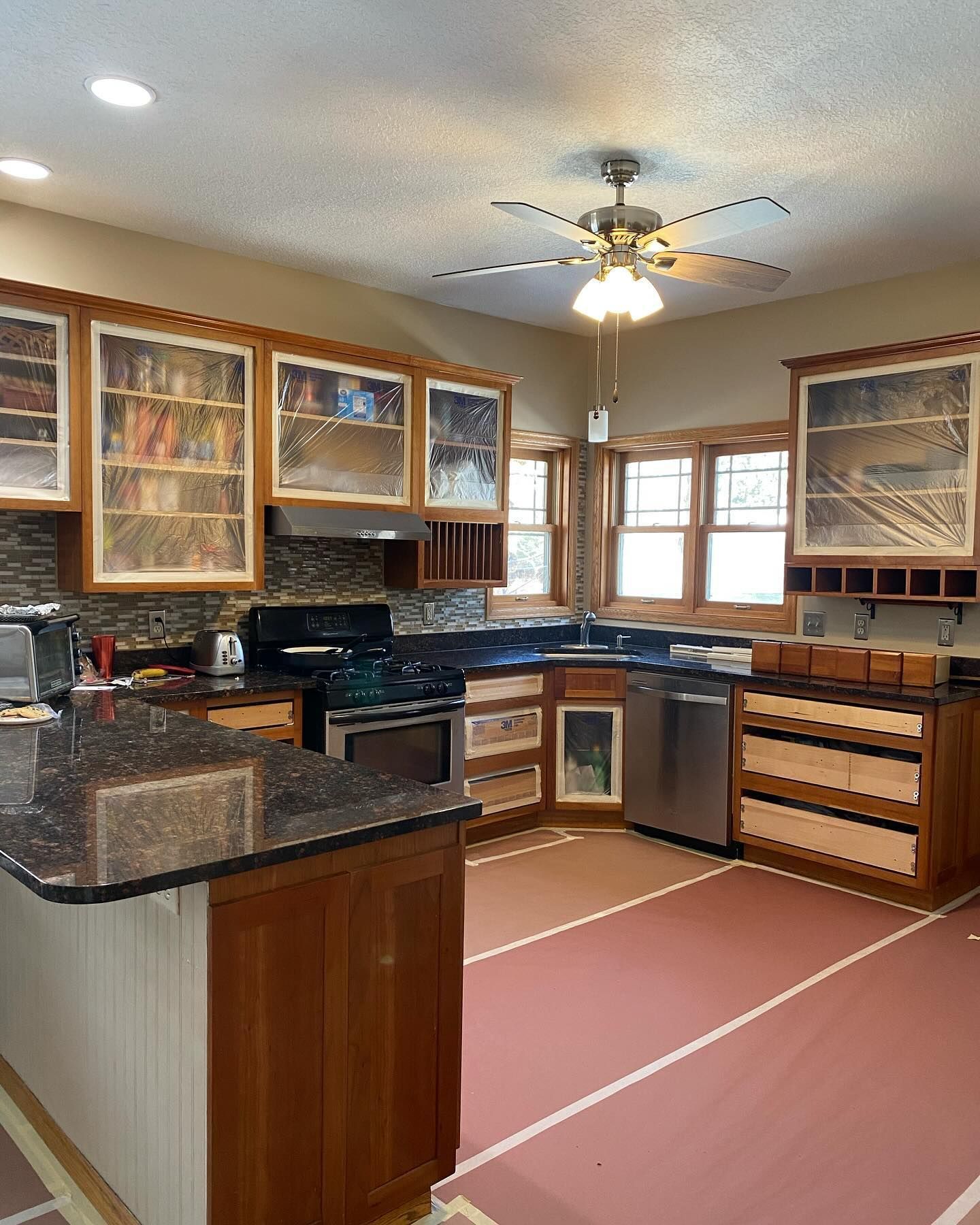 Kitchen under renovation: wooden cabinets, stainless steel appliances, dark countertops, and reddish flooring with masking tape lines.