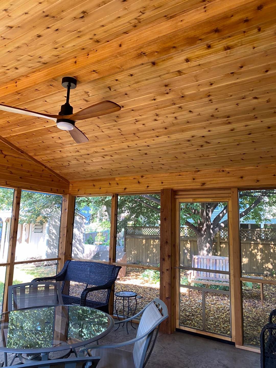 A screened porch with a wooden ceiling and a ceiling fan. It has seating and views of a backyard.