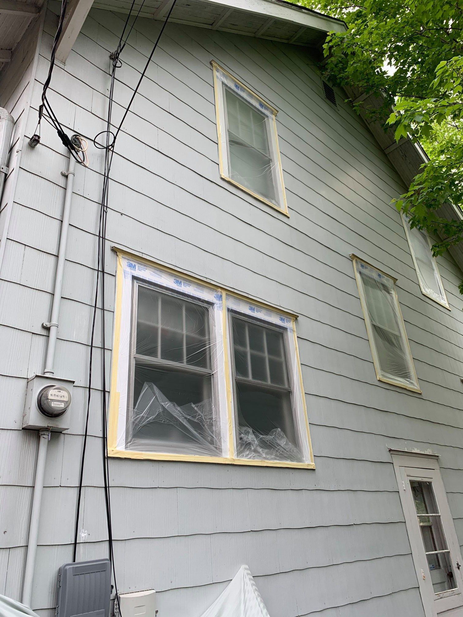 Gray house exterior with multiple windows covered in plastic and yellow tape. Electrical wires and meter are visible.