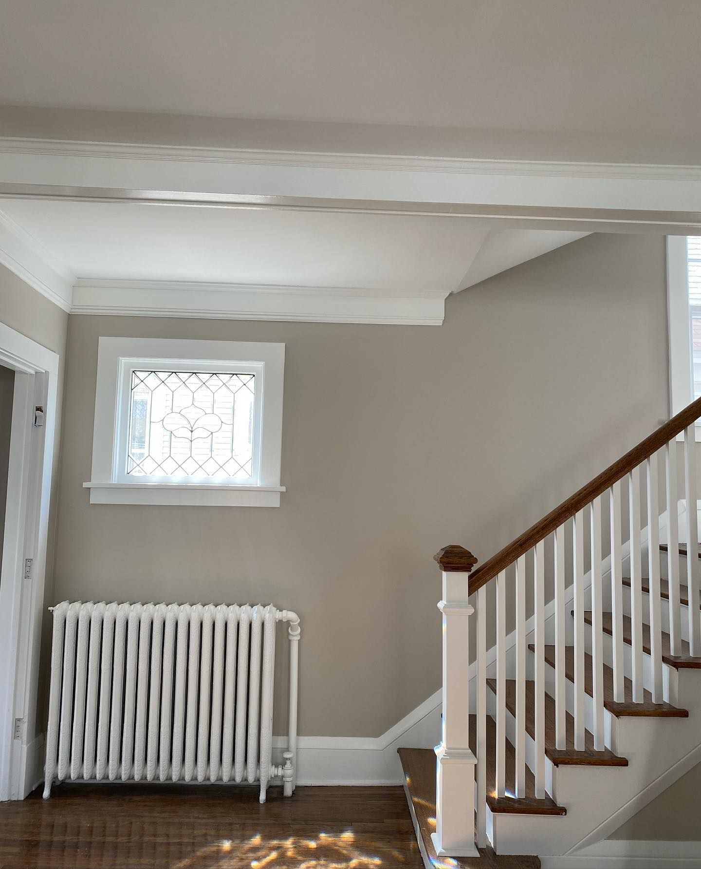 Entryway with beige walls, white trim, and a wooden staircase. A radiator sits below a small, leaded-glass window.