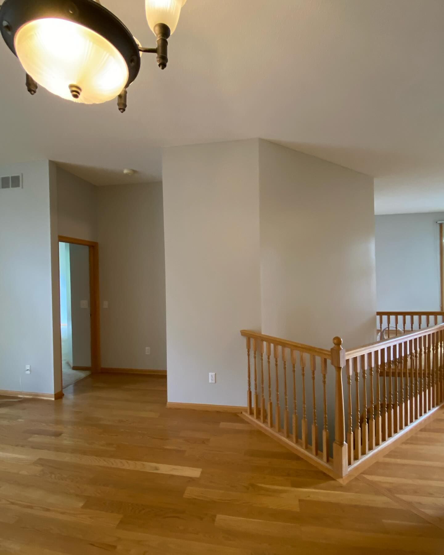 Interior view of a home with light wooden floors, beige walls, a wooden banister, and a hallway with a doorway. A light fixture hangs from the ceiling.