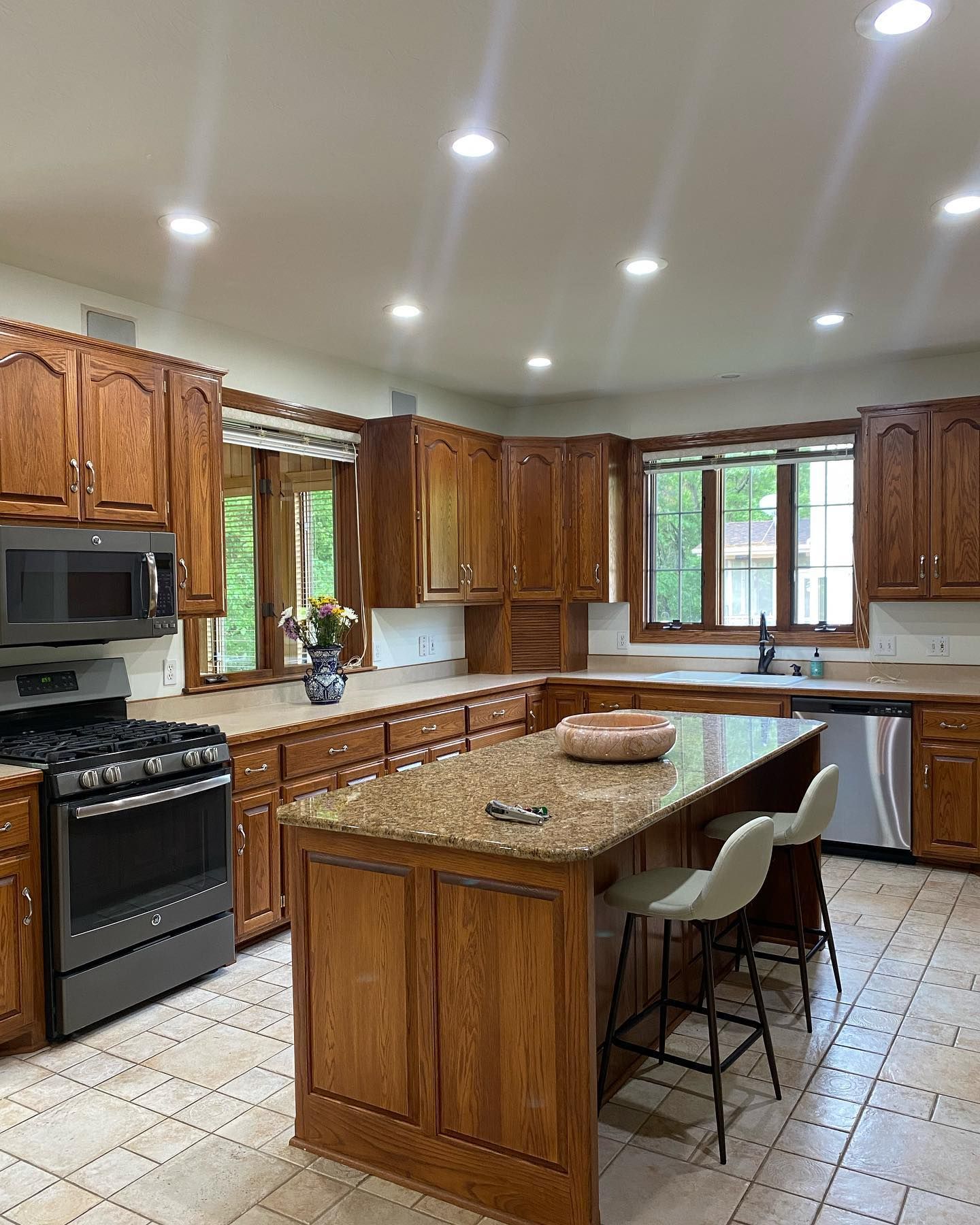 Spacious kitchen with wood cabinets, granite countertops, and a central island with seating. Stainless steel appliances and light-colored tile floors are present.