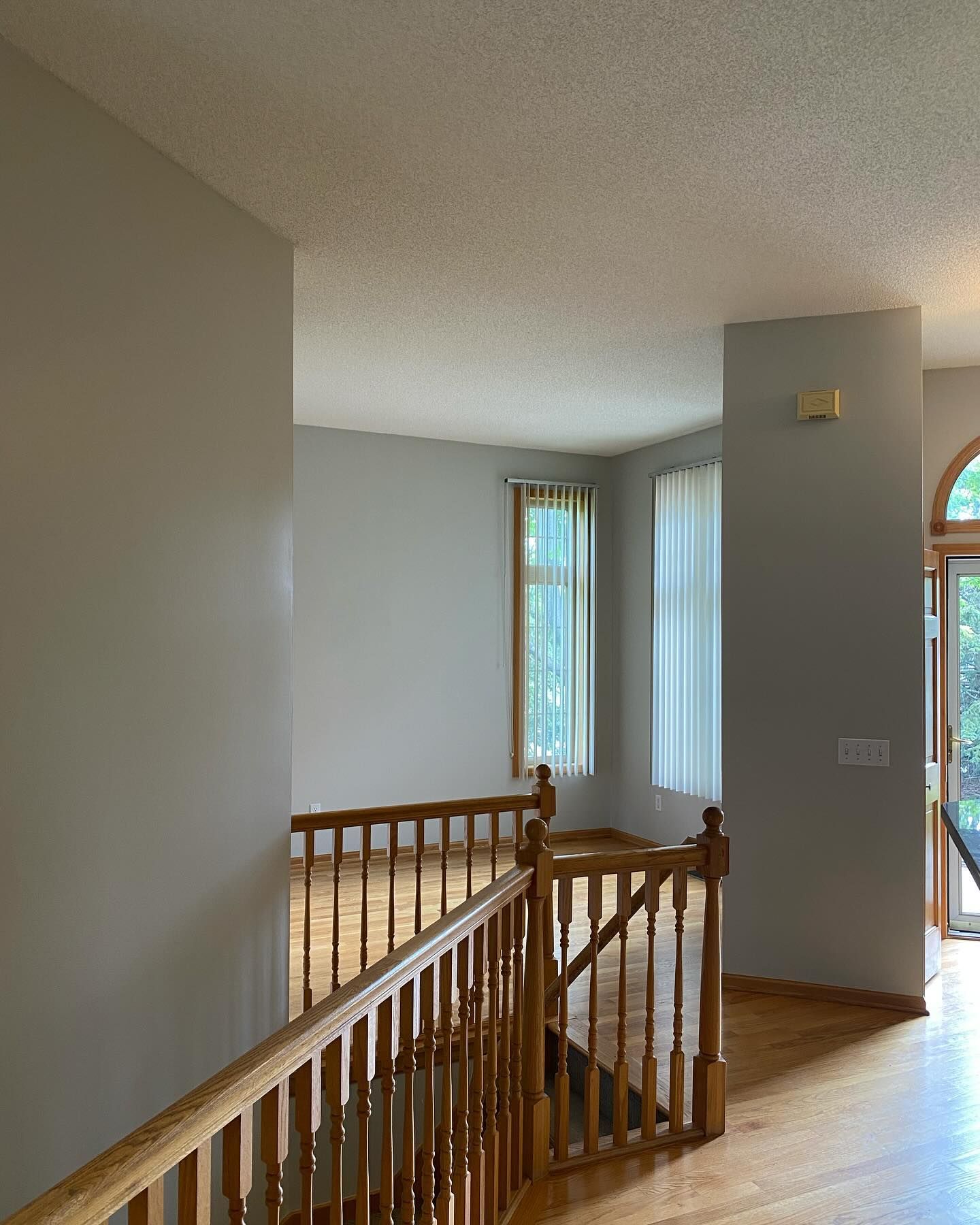 Interior view from a home's second floor, showing a wooden banister, light gray walls, hardwood floors, and a room with windows and white curtains.