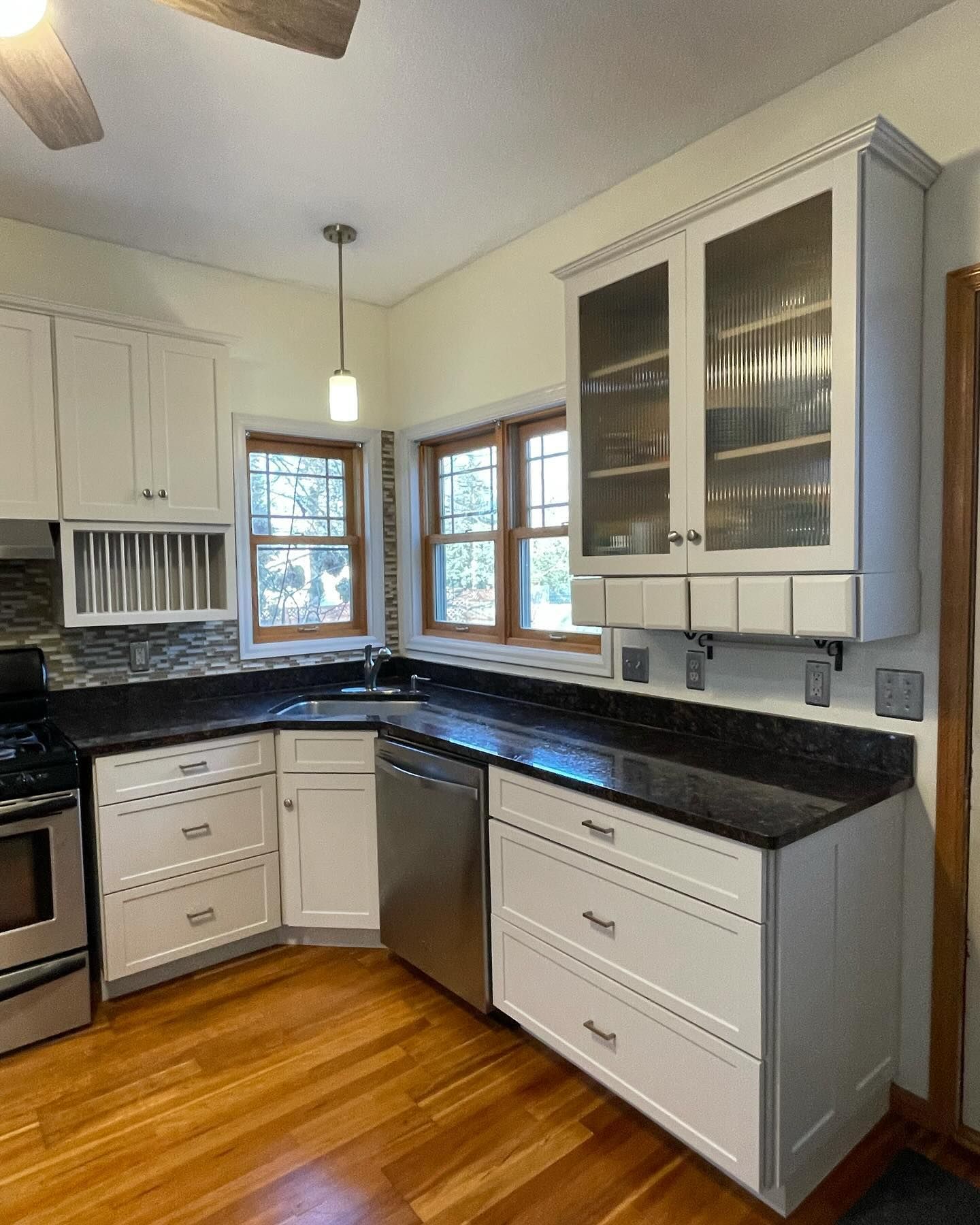White kitchen with black countertops, stainless steel appliances, and glass-front upper cabinets. Natural light streams in through the windows.
