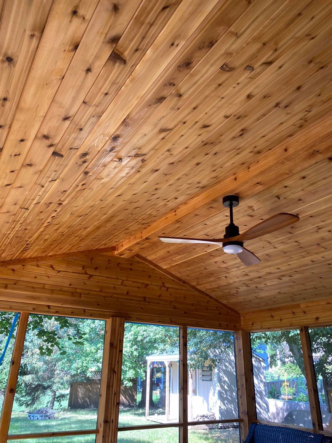 Cedar wood ceiling of a screened porch with a ceiling fan. View through screens to a yard with trees and a shed.