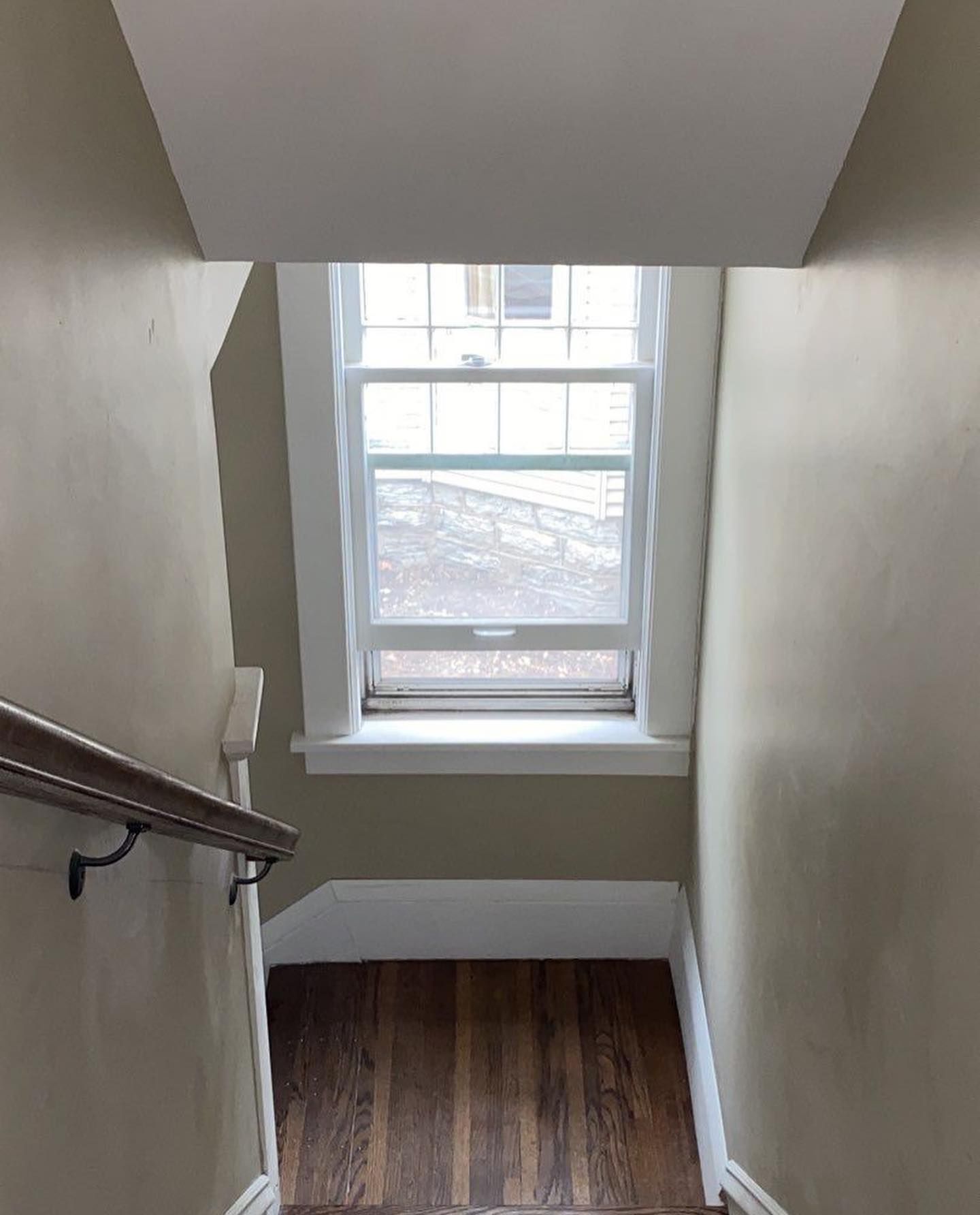 Looking down a staircase toward a window. White window frame against the beige walls and a dark wooden floor.