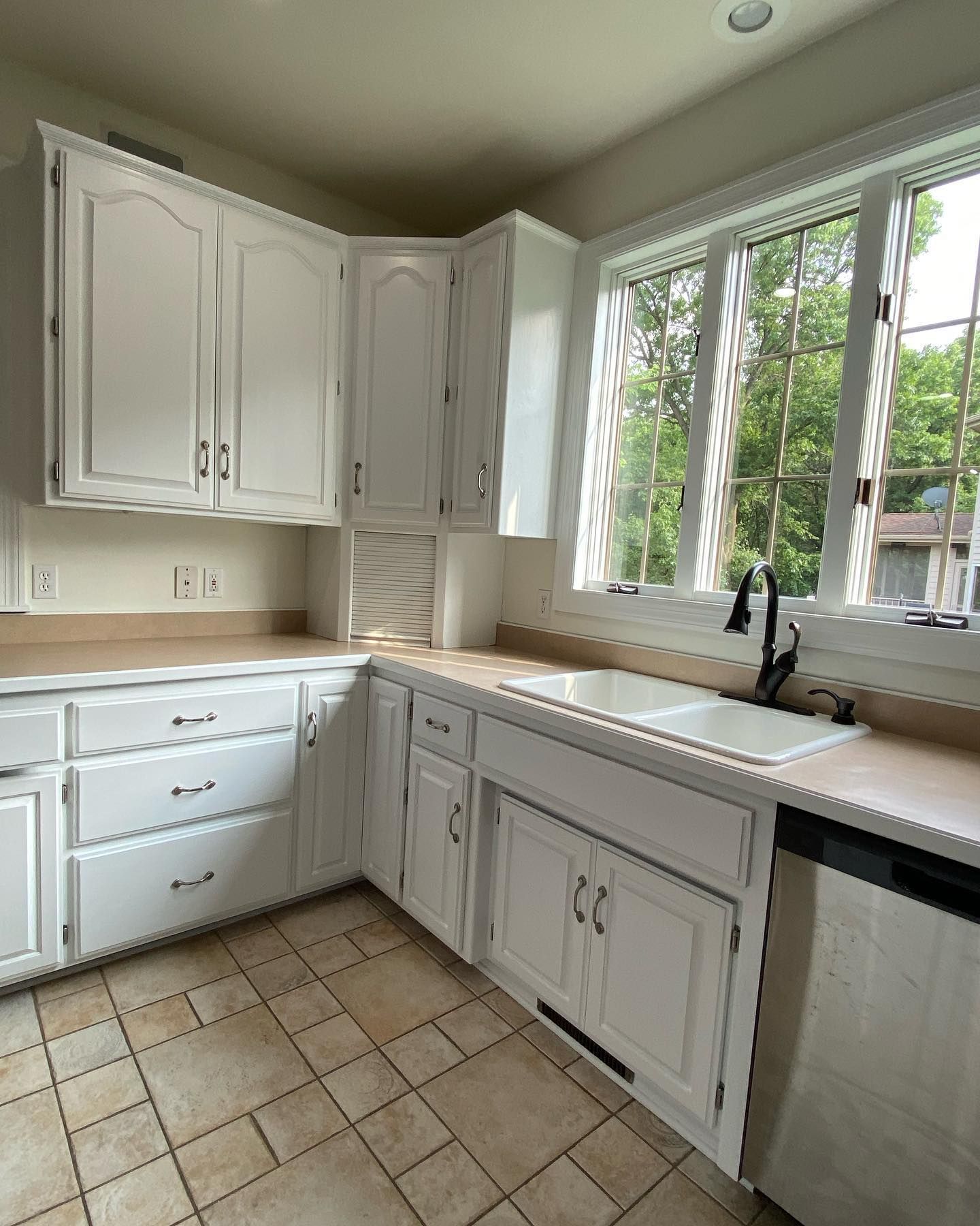 Corner kitchen with white cabinets, tan countertops, and tile floor. A sink sits under a large window.