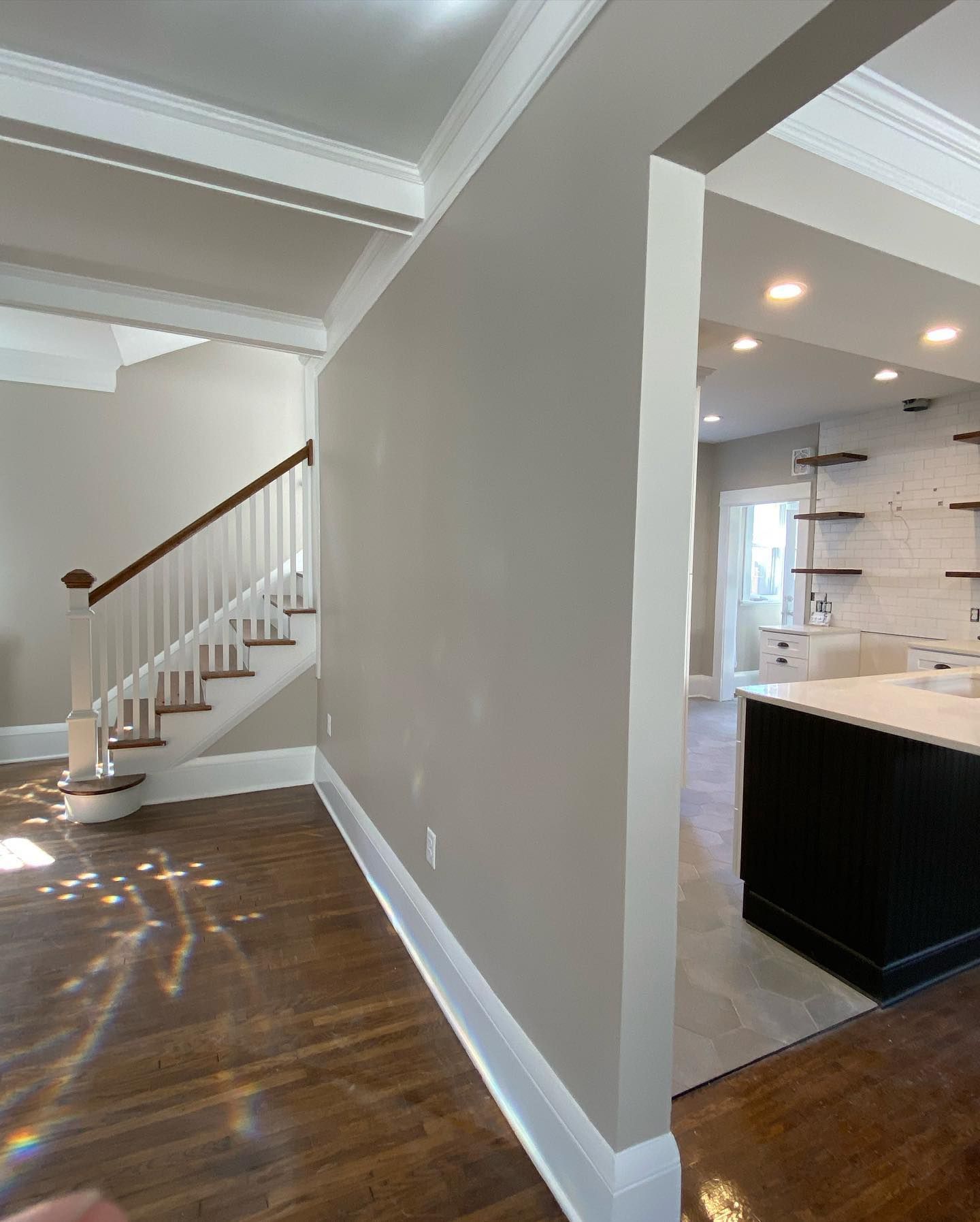 A light-filled interior with a staircase, a neutral-colored wall, and a glimpse of a kitchen with a black island and shelving. Wooden floors.