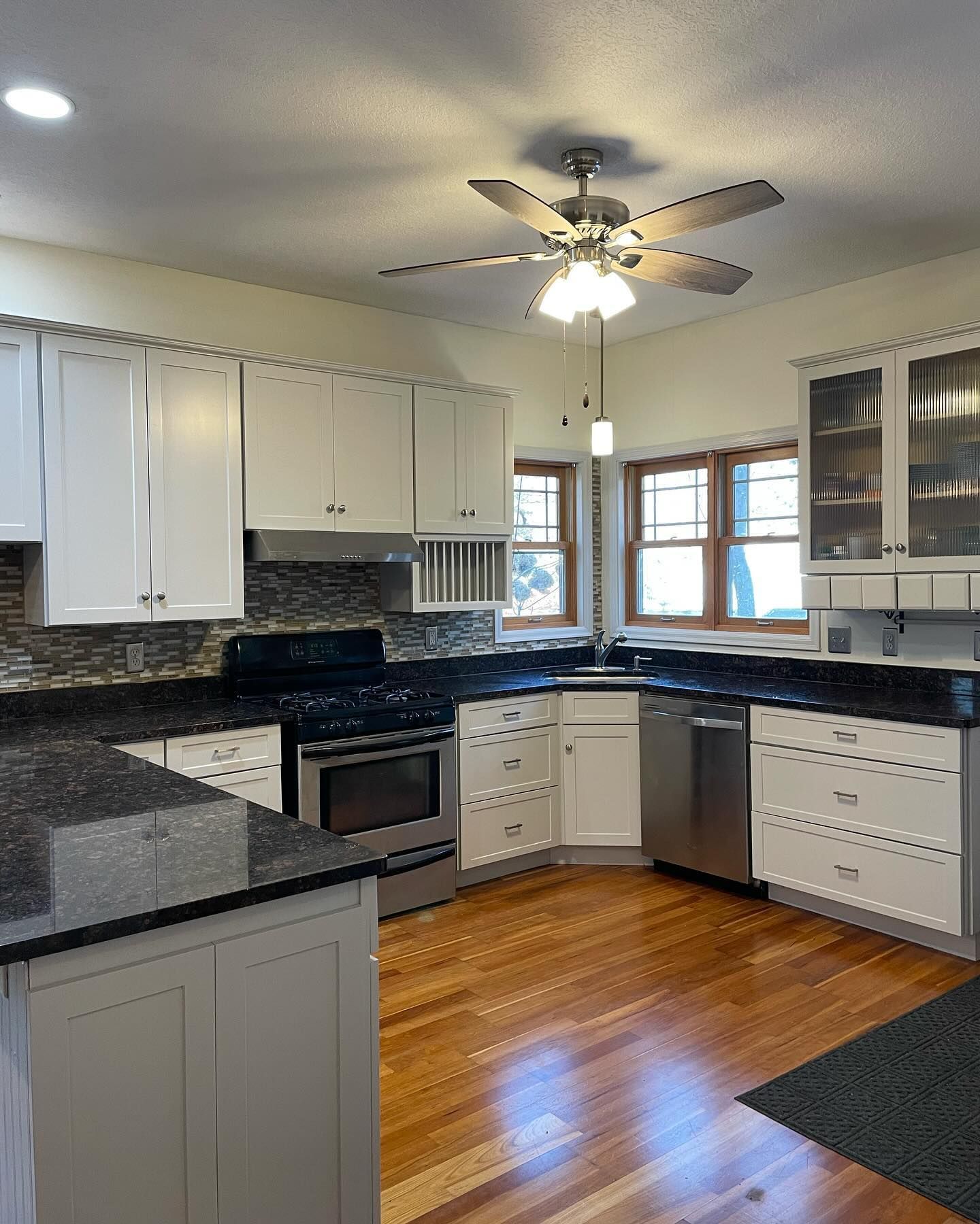 A well-lit kitchen with white cabinets, dark countertops, stainless steel appliances, and hardwood floors. A ceiling fan hangs overhead.