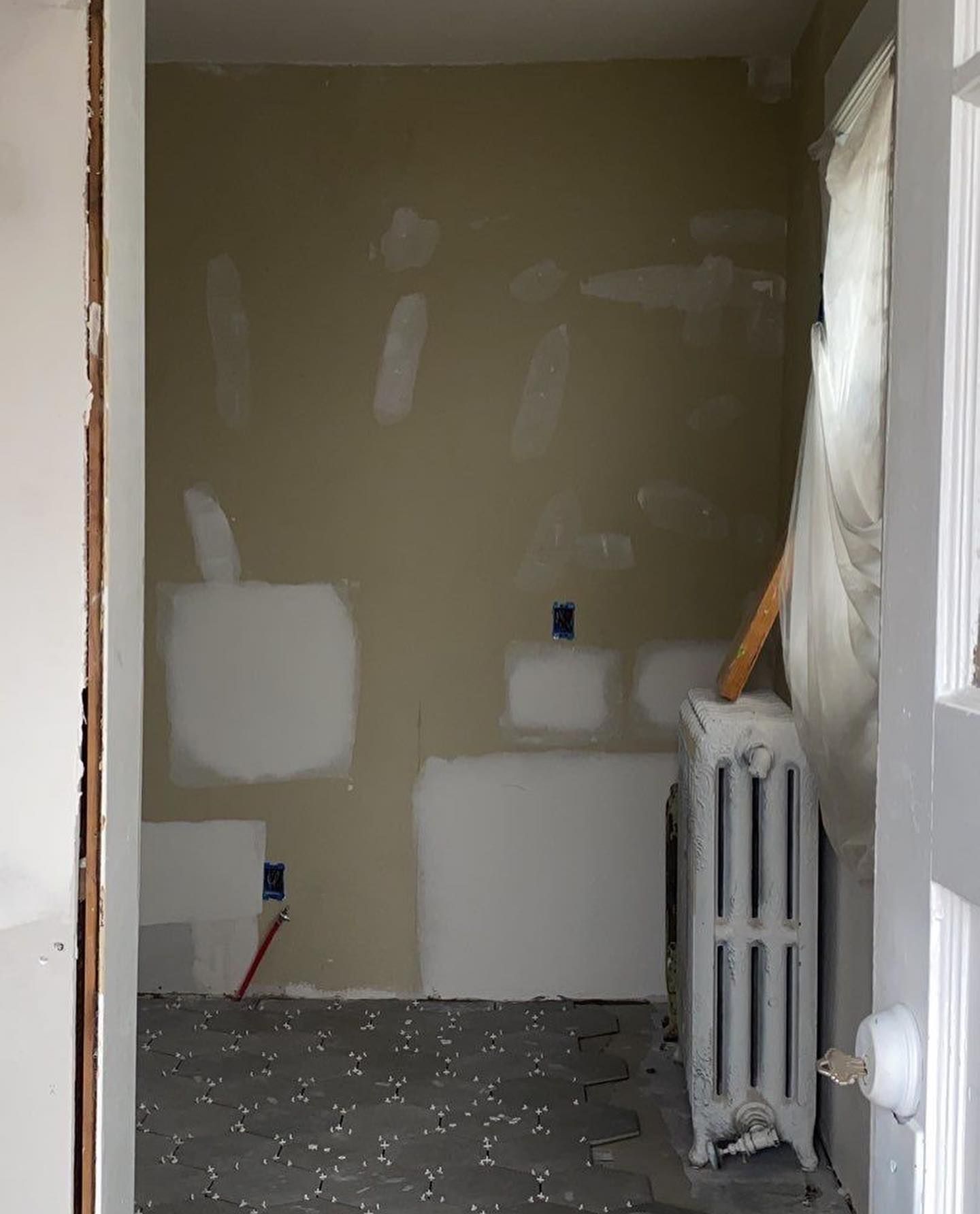 Empty room under renovation with tan walls, gray floor, and a radiator. Patches of drywall are visible.