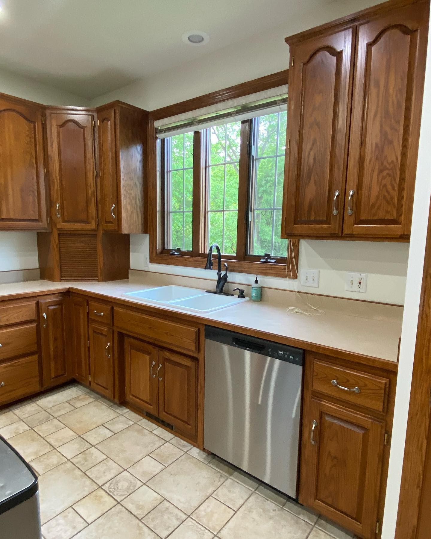 Kitchen with medium-tone wood cabinets, light countertops, a stainless steel dishwasher, and a sink in front of a window with a green view.