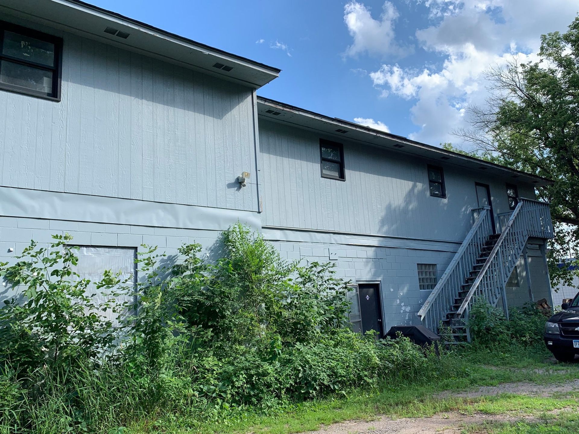 Two-story apartment building with blue siding and a wooden staircase. Overgrown vegetation covers the building's base.