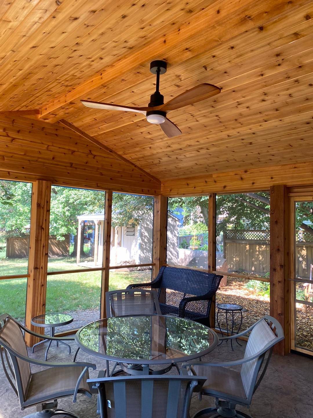 Screened porch with wood ceiling, ceiling fan, and outdoor furniture.  View through screens to yard with trees and a shed.