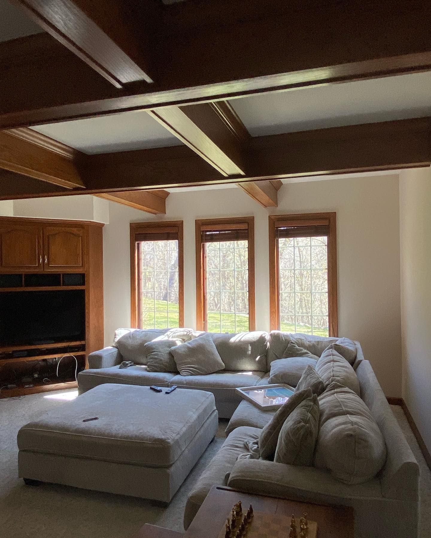 Living room with beige sectional sofa, ottoman, and built-in entertainment center. Sunlight streams through windows. Brown wooden beams cross the ceiling.