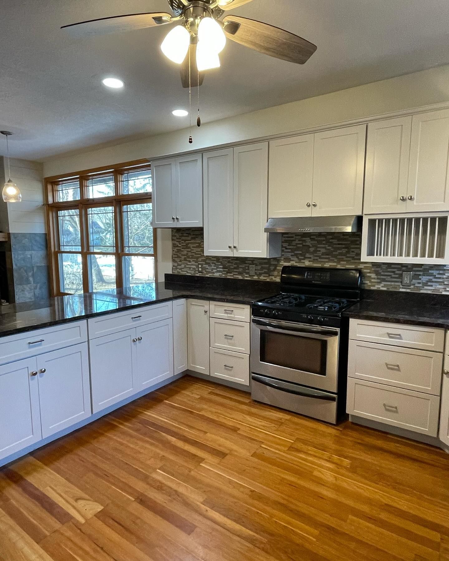 A kitchen with white cabinets, dark countertops, a stainless steel oven, and wood flooring. A window offers an outdoor view.