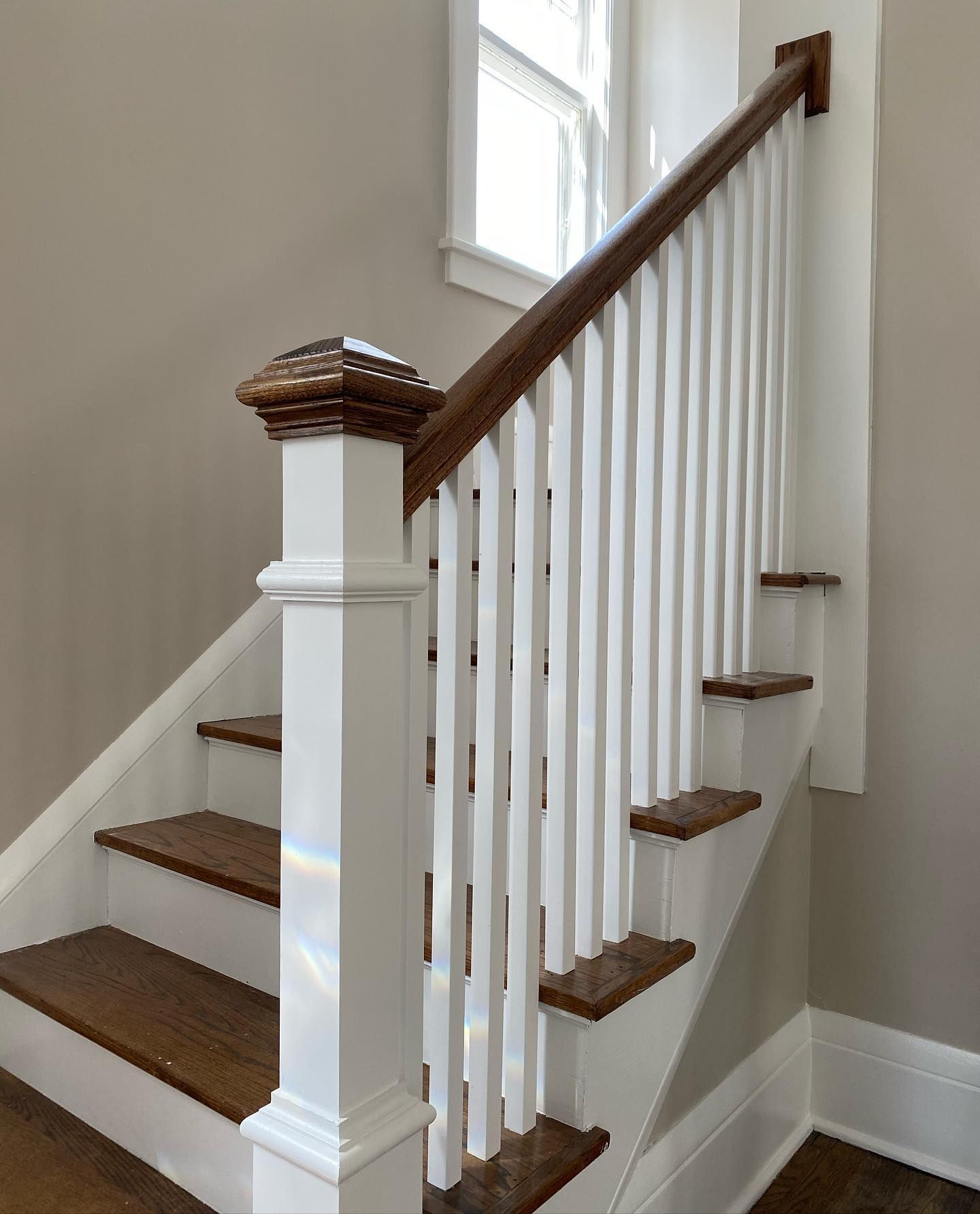 Staircase with white balusters, brown steps, and a dark wood handrail and newel post. Natural light streams through the window.