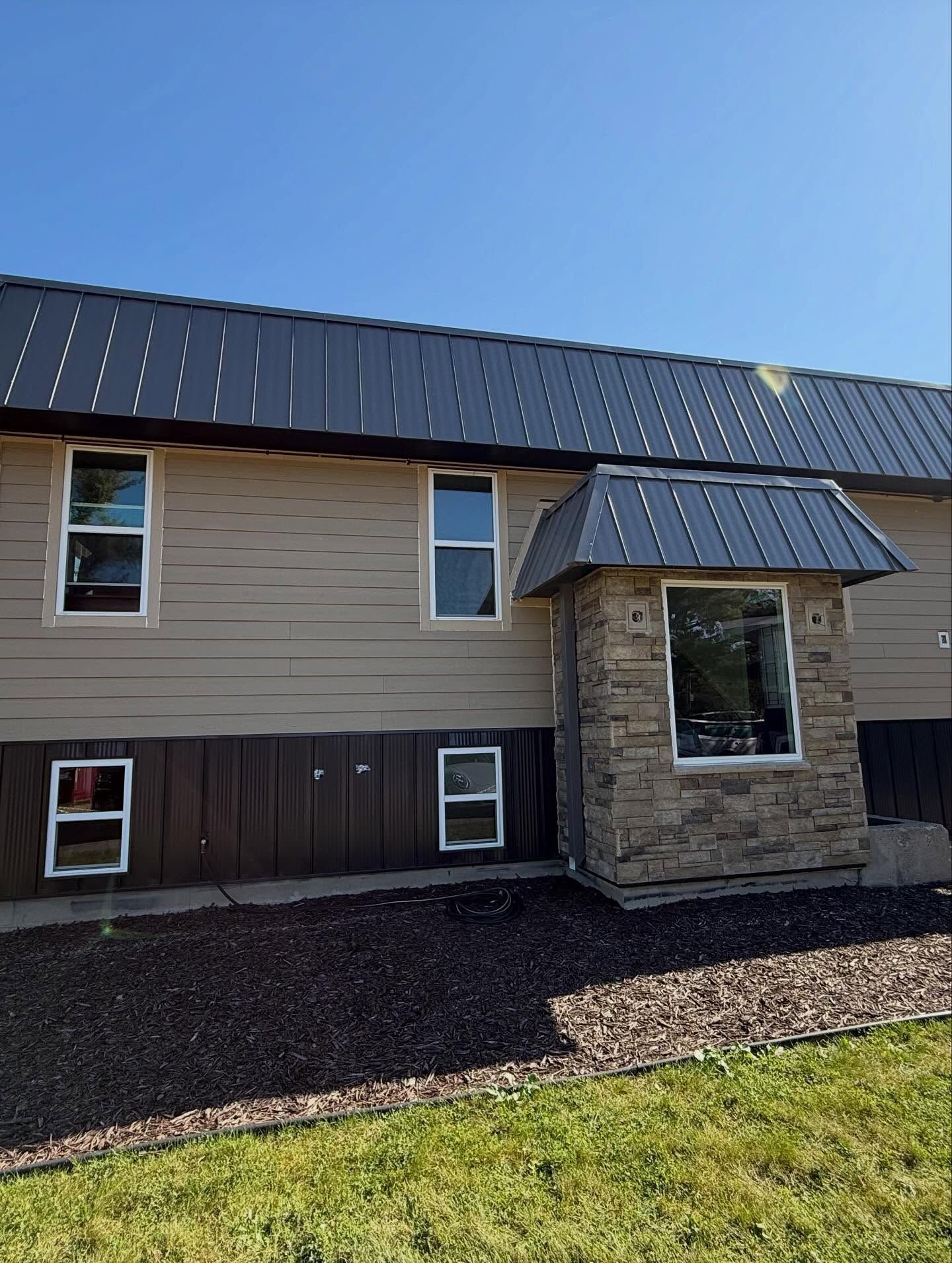 Tan house with dark metal roof, stone entryway, windows. Brown siding and lower trim. Sunny day.