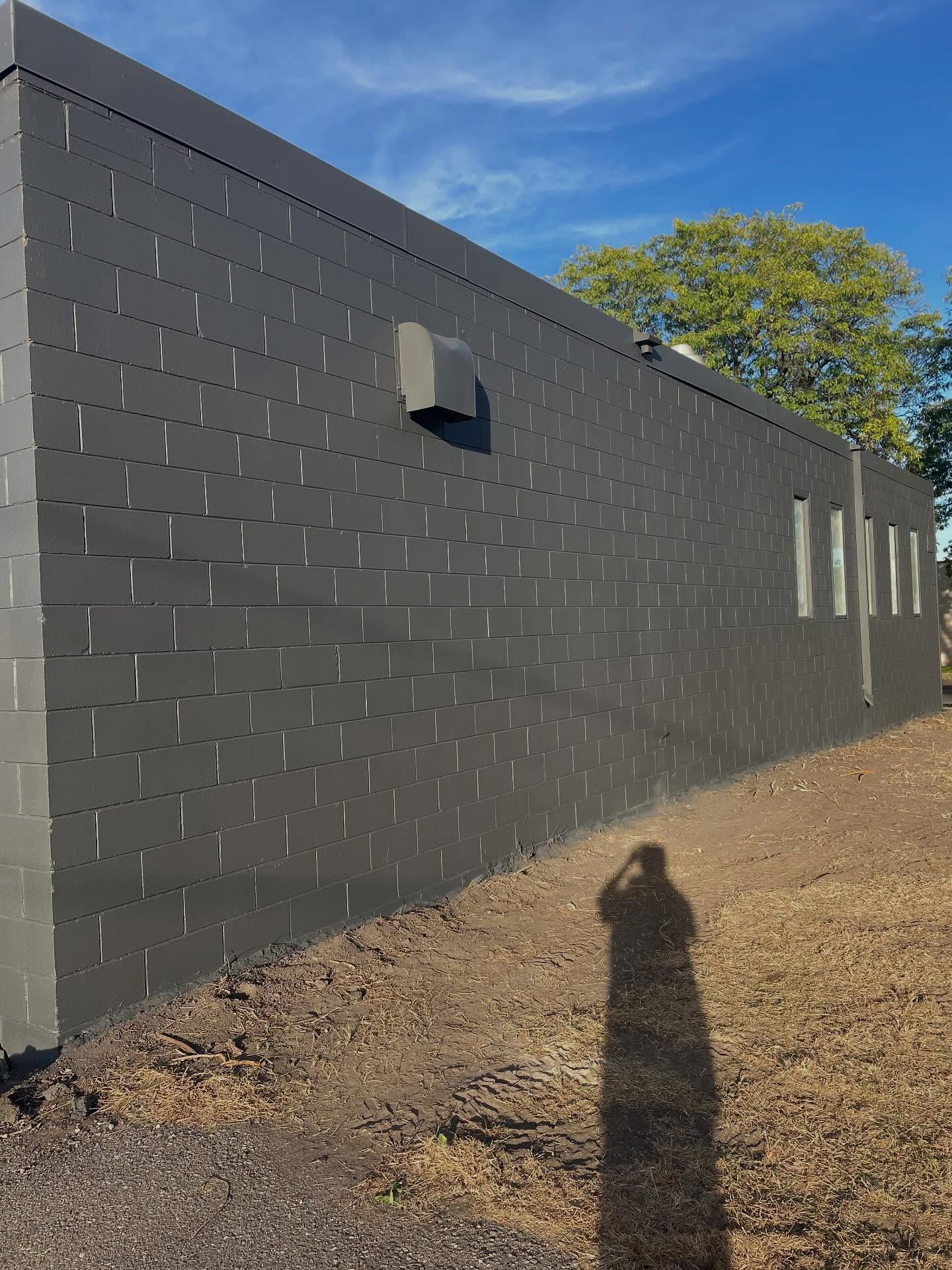 Dark gray building with a rectangular air vent, several small windows, and a long shadow on a sunny day.