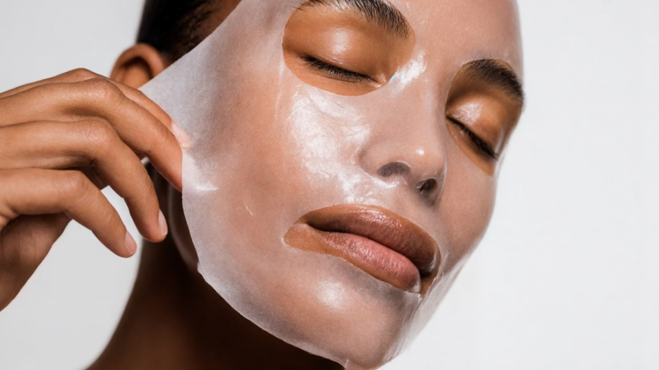 Person removing a sheet mask from their face. Eyes are closed, skin is glowing.