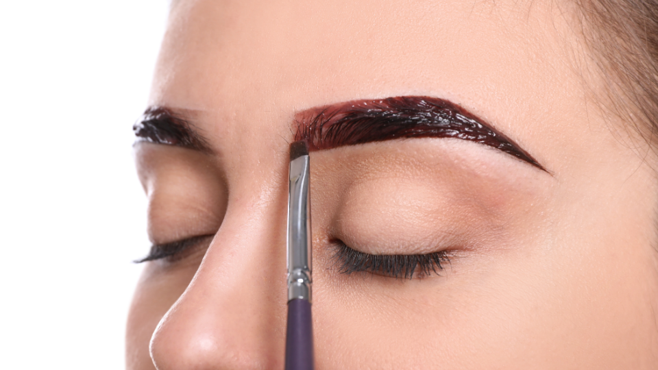 A person's closed eye as a brush applies dark eyebrow dye to one eyebrow, white background.