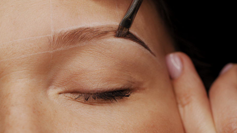 Close-up of a person's eye, brow being shaped with a brush. Dark brown pigment is applied.