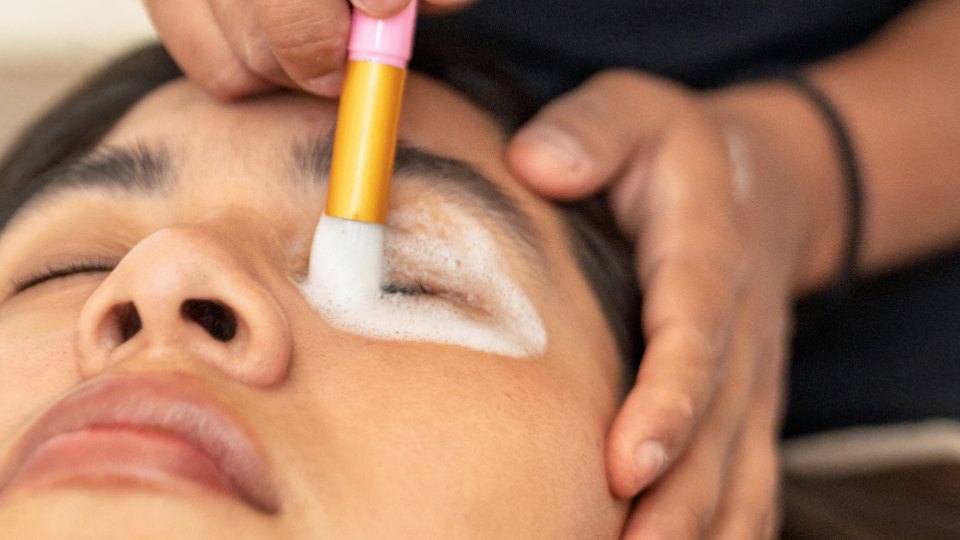 Person's eye being cleaned with a brush. The person is lying down with closed eyes. A hand is holding the brush.