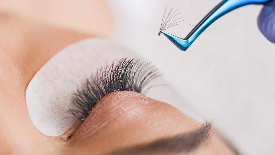 Eyelash extensions being applied; tweezers holding a fan of lashes near a closed eye with a protective pad.