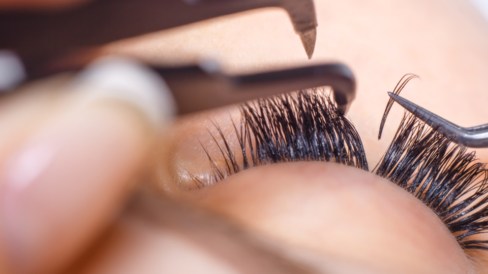 Close-up of a person's eye with lash extensions being applied with tweezers.