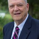 Man in navy suit, white shirt, and pink-striped tie smiles outdoors.